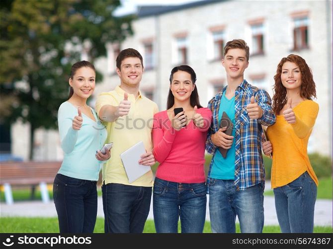 friendship, technology, education, school and people concept - group of smiling teenagers with smartphones and tablet pc computers showing thumbs up over campus background