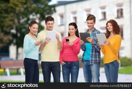 friendship, technology, education, school and people concept - group of smiling teenagers with smartphones and tablet pc computers over campus background