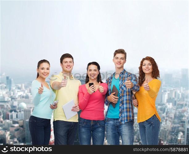 friendship, technology, gesture and people concept - group of smiling teenagers with smartphones and tablet pc computers showing thumbs up over city background