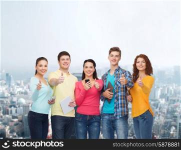friendship, technology, gesture and people concept - group of smiling teenagers with smartphones and tablet pc computers showing thumbs up over city background