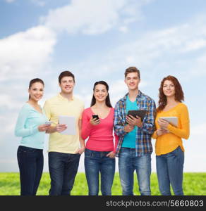 friendship, technology, summer and people concept - group of smiling teenagers with smartphones and tablet pc computers over blue sky and grass background