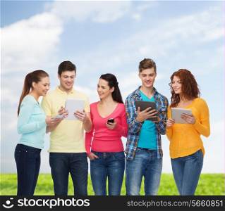 friendship, technology, summer and people concept - group of smiling teenagers with smartphones and tablet pc computers over blue sky and grass background
