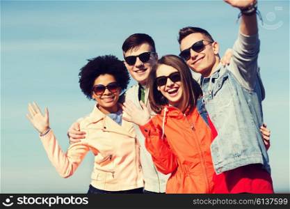 friendship, tourism, travel and people concept - group of happy teenage friends in sunglasses hugging and waving hands outdoors