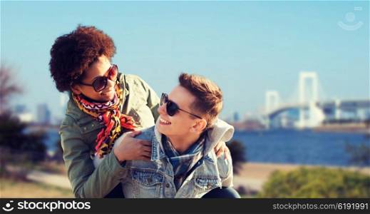 friendship, travel, tourism and people concept - happy international teenage couple in shades having fun over rainbow bridge at tokyo in japan background