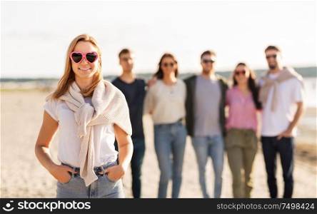 friendship, valentine&rsquo;s day and people concept - happy woman in heart-shaped sunglasses with group of friends on beach in summer. happy woman with friends on beach in summer