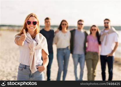friendship, valentine&rsquo;s day and people concept - happy woman in heart-shaped sunglasses with group of friends on beach in summer showing thumbs up. woman with friends on beach showing thumbs up