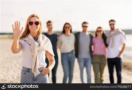 friendship, valentine&rsquo;s day and people concept - happy woman in heart-shaped sunglasses with group of friends on beach in summer waving hand. woman with friends on beach in summer waving hand