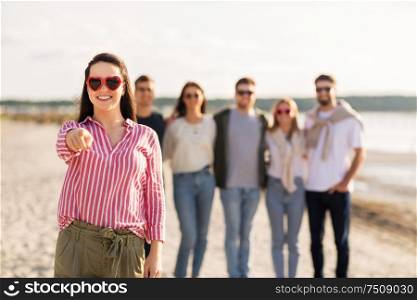 friendship, valentine&rsquo;s day and people concept - happy woman in heart-shaped sunglasses with group of friends on beach in summer pointing to you. woman with friends on beach pointing to you