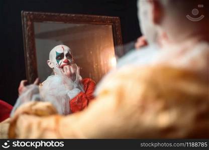 Frightened bloody clown with crazy eyes looking at the mirror, nightmare. Man with makeup in carnival costume, mad maniac