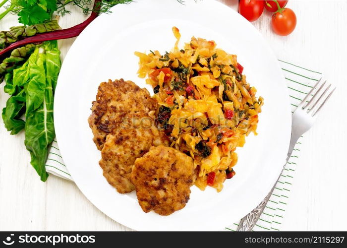 Fritters of minced meat with stewed cabbage in a plate, fork on towel, tomatoes, parsley and chard on wooden board background from above