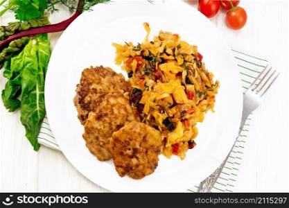 Fritters of minced meat with stewed cabbage in a plate, fork on towel, tomatoes, parsley and chard on wooden board background from above