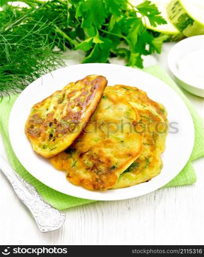Fritters of zucchini, dill and parsley in a plate on a towel, sour cream in saucer on the background of light wooden board