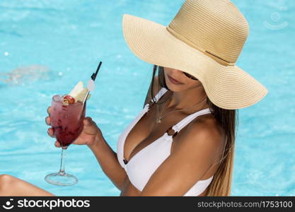 From above of anonymous woman in white bikini and straw hat sitting on poolside with turquoise water holding cocktail. Young woman with cocktail on poolside