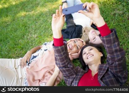 From above of delighted multiracial female friends taking self portrait on smartphone while lying on grassy lawn on summer day. Cheerful diverse women taking selfie on lawn