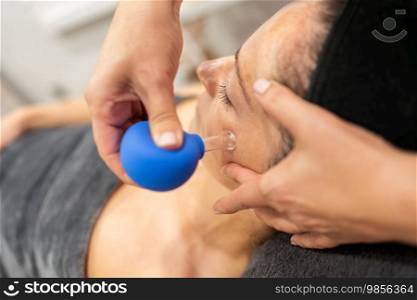 From above of hand applying cupping therapy with blue facial cup to woman with dark hair during skin treatment in beauty spa. Client receiving facial cupping in beauty parlor