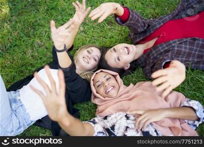 From above of happy young multiracial women smiling and waving hands at camera while lying on grassy meadow in park. Cheerful young diverse female friends lying on grass and smiling at camera