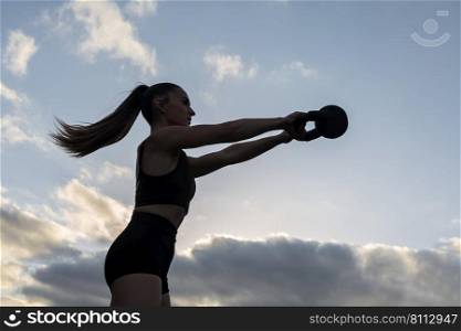 From below fit female athlete in sportswear with ponytail lifting kettlebell in outstretched arms against cloudy sunrise sky during morning fitness workout. Sportswoman exercising with kettlebell at sunrise