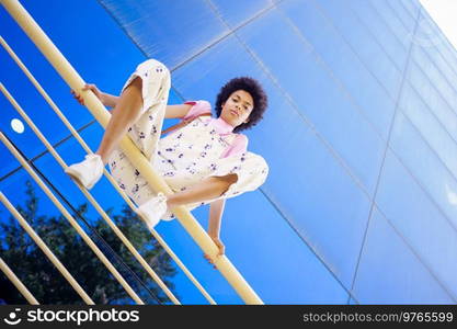 From below full body of African American female looking at camera while sitting on fence near glass building in city. Serious black woman sitting on railing