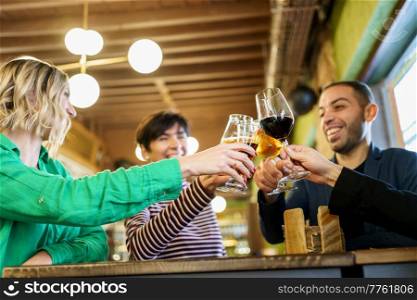 From below multiracial man and women smiling and clinking glasses of alcohol beverages over table while proposing toast during meeting in bar. Diverse friends clinking glasses over table in beautiful pub