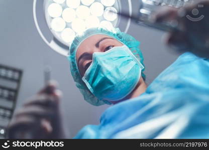 From below woman surgeon in medical uniform using professional tools while standing under bright light in operating theater. High quality photo. From below woman surgeon in medical uniform using professional tools while standing under bright light in operating theater