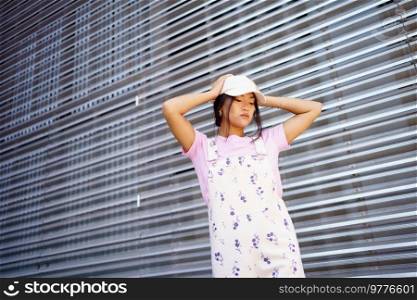 From below young Asian female in stylish clothes and cap touching head and looking down while standing near metal wall of modern building. Stylish Asian woman near metal wall