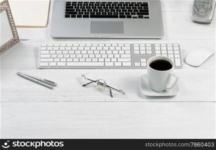 Front view angle of desktop layout for proper use consisting of laptop, keyboard, pens, mouse, picture frame, phone, coffee, reading glasses and work folders on white desk.