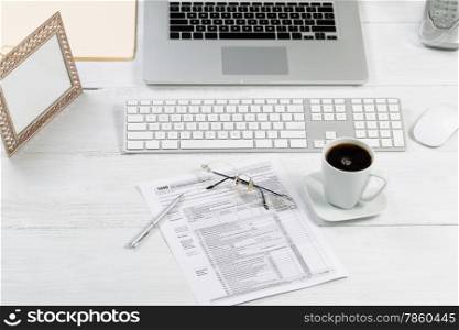 Front view angle of desktop layout for proper use consisting of laptop, keyboard, pens, mouse, picture frame, phone, coffee, reading glasses, tax forms and work folders on white desk.
