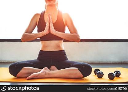 Front view beauty woman doing yoga and raise hand or pay obeisance in fitness workouts training gym center. Lifestyle sport woman sitting on mat with sport equipment and exercise dumbbells background