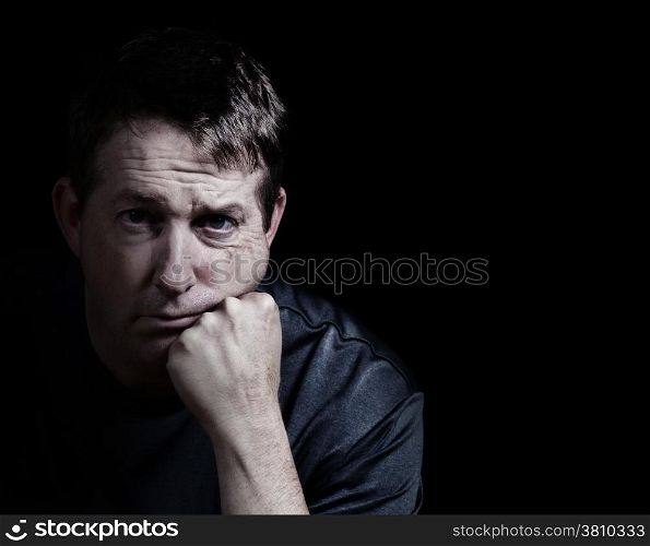 Front view close up of mature man looking forward with his chin in hand displaying depression on black background