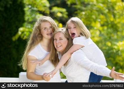 Front view, looking forward, of mother holding her younger daughter with older daughter hugging them while outdoors on patio with blurred out woods in background