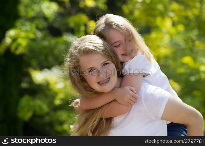 Front view, looking forward, with focus on older sister carrying younger sister on her back while outdoors on patio with blurred out trees in background