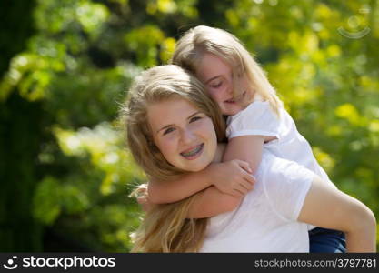 Front view, looking forward, with focus on older sister carrying younger sister on her back while outdoors on patio with blurred out trees in background