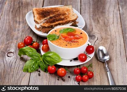 Front view of a bowl of fresh creamy tomato soup, tuna sandwich, with cherry tomatoes and basil leaves on rustic wooden boards.