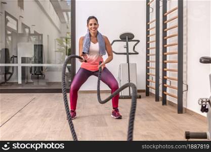 Front view of a cheerful young adult woman in sportswear exercising with battle ropes at the gym.. Young adult woman in sportswear exercising with battle ropes at the gym