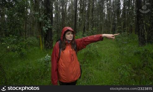 Front view of a Hispanic woman with a black backpack and a red waterproof jacket pointing the direction with her outstretched arm in the middle of a forest during the day. Front view of a Hispanic woman with a black backpack and a red waterproof jacket pointing the direction with her outstretched arm