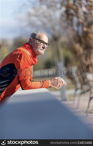 Front view of a senior runner male with sportswear sitting on a wooden fence while looking away