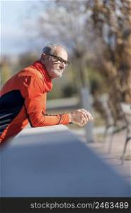 Front view of a senior runner male with sportswear sitting on a wooden fence while looking away