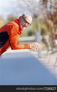 Front view of a senior runner man with sportswear sitting on a wooden fence while looking camera