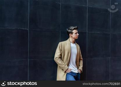 Front view of a trendy young man standing against black wall while looking away outdoors in the street