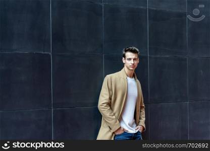 Front view of a trendy young man standing against black wall while looking camera outdoors in the street