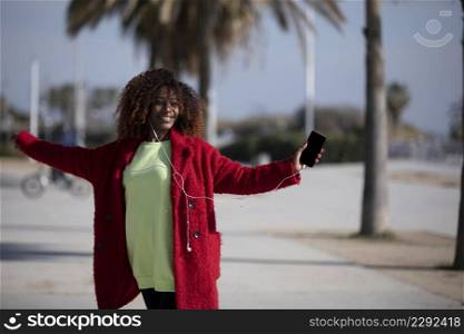 Front view of a young beautiful curly african american woman jumping and dancing while listening music and smiling outside
