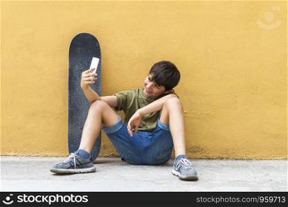 Front view of a young boy sitting on ground leaning on a yellow wall while using a mobile phone to take a selfie