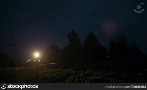 Front view of an illuminated train passing fast through a rural area, by night. Raining