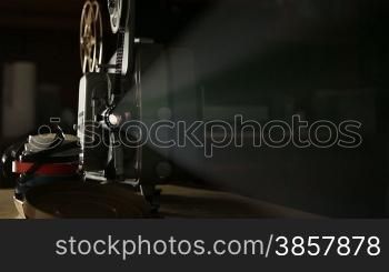 Front view of an old-fashioned antique Super 8mm film projector, projecting a beam of light in a dark room next to a stack of unraveled film reels. Includes projector audio.