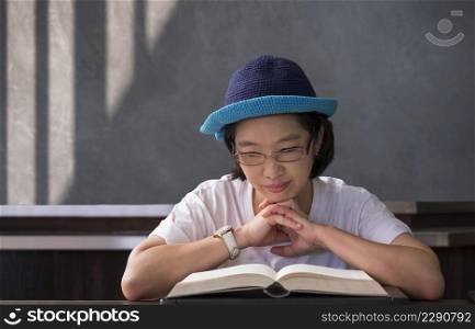 Front view of Asian casual woman in eyeglasses reading book on wooden table in vintage living room