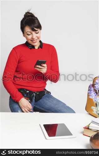 Front view of beautiful latin woman sitting on a stool while using a mobile phone at office against white background