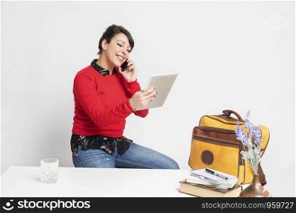Front view of beautiful woman with ponytail sitting on a stool while using phone and holding a tablet pc against white background
