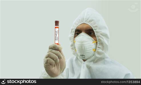 Front view of doctor with mask and bioprotective suit holding a Covid-19 labeled blood sample test tube with his hand and staring intently at it on white background. Doctor with mask and bioprotective suit holding a Covid-19 labeled blood sample test tube with his hand and staring intently at it on white background