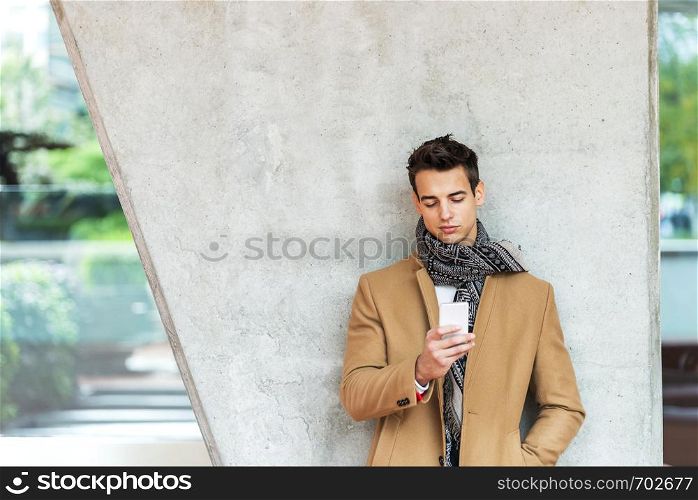 Front view of fashionable young man wearing denim clothes leaning on a wall while using a mobile phone outdoors