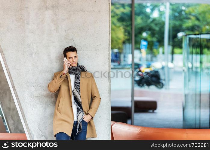 Front view of fashionable young man wearing denim clothes leaning on a wall while using a mobile phone outdoors